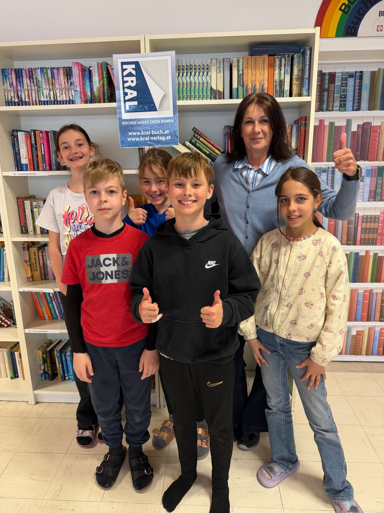 A woman and five children stand in front of a bookshelf, smiling and giving thumbs up. The woman wears a light blue cardigan.