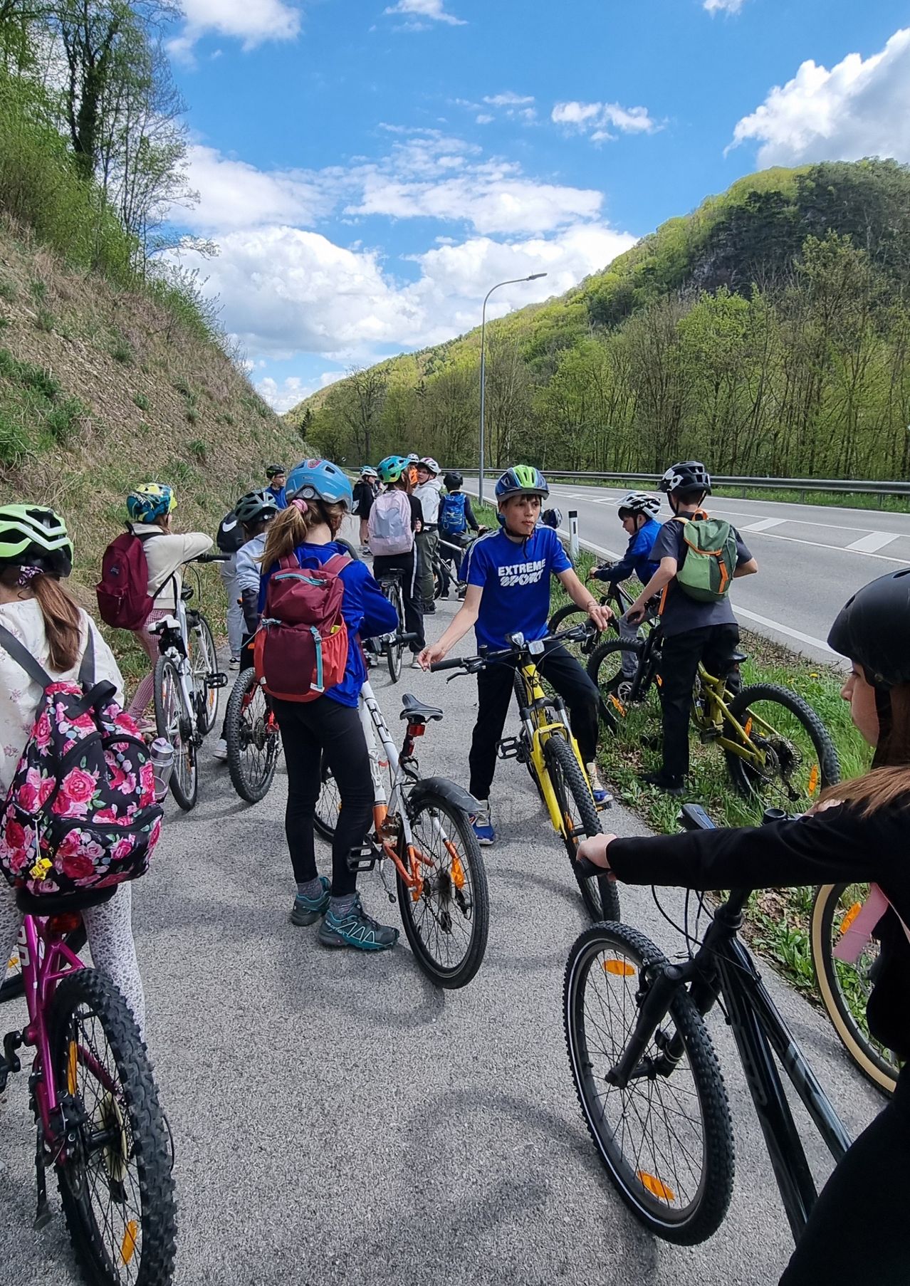 A group of cyclists with helmets on a road, some preparing to start a ride. They have backpacks and various colors of bicycles. A hill with greenery is on one side.