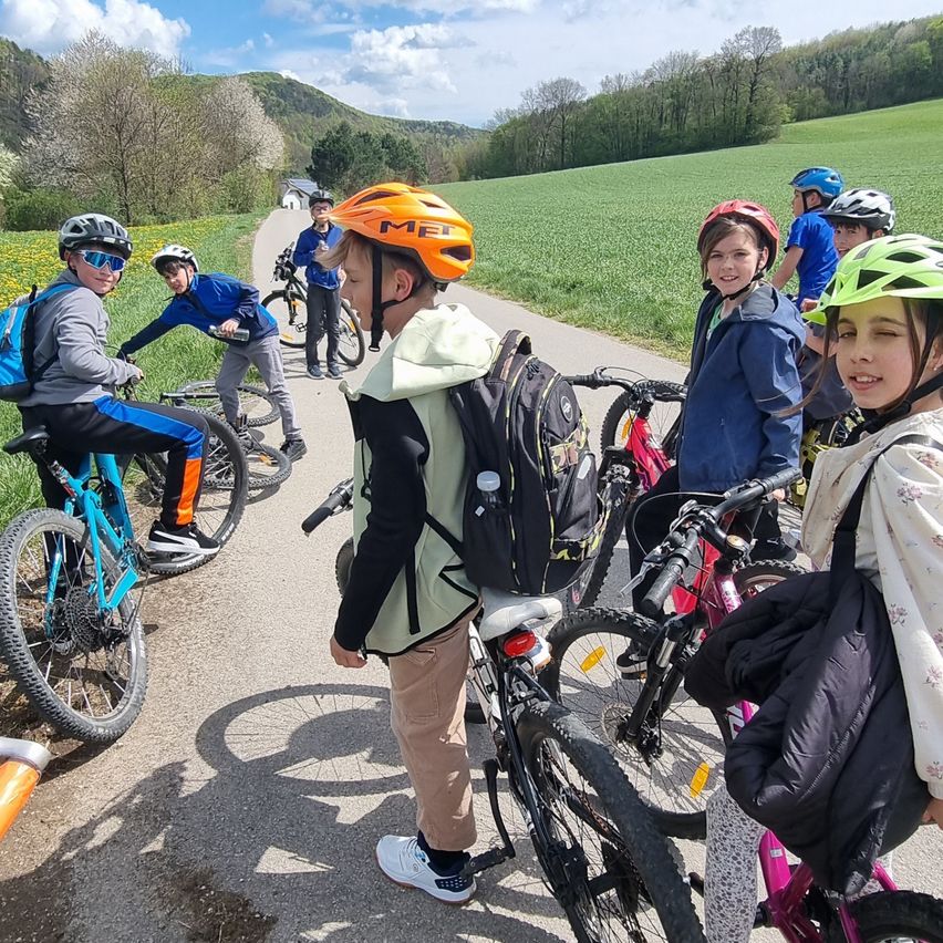 A group of kids on bicycles, wearing helmets, are gathered on a pathway in a grassy field. They are smiling, some holding their bikes, others standing around. The weather is sunny.