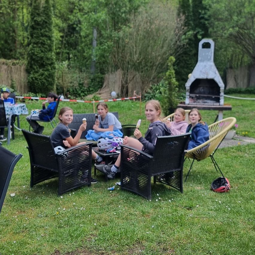 A group of kids are sitting on chairs around a table, eating ice cream. Some are wearing helmets. Behind them is a grill, and there are trees in the background.