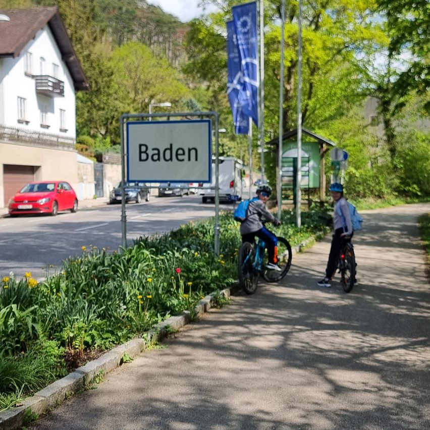Two cyclists with helmets and backpacks are on the side of a road in Baden. A house and cars are in the background.