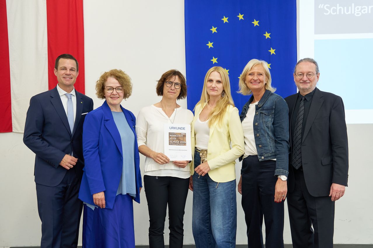 Five people pose for a photo, three women and two men, with one woman holding a certificate. Behind them is a backdrop with a European Union flag and a red flag.