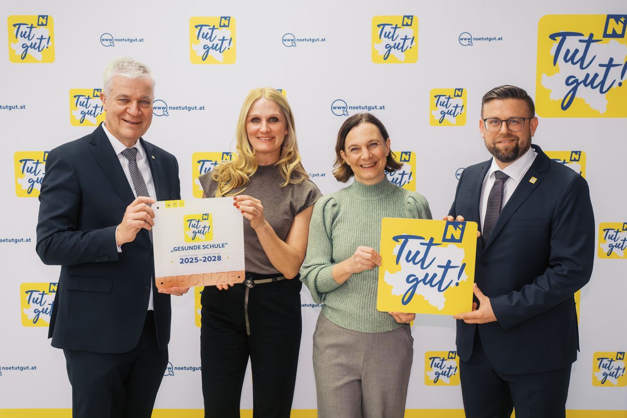 Vier Personen stehen vor einer Wand mit einem gelben Schild, das 'Tuit gut!' auf Deutsch sagt. Sie halten gelbe Schilder mit 'Tuit gut!' und 'GESUNDE SCHUL'. Der Mann auf der linken Seite hält ein weißes Schild mit 'SPONSOR'. Sie alle lächeln.