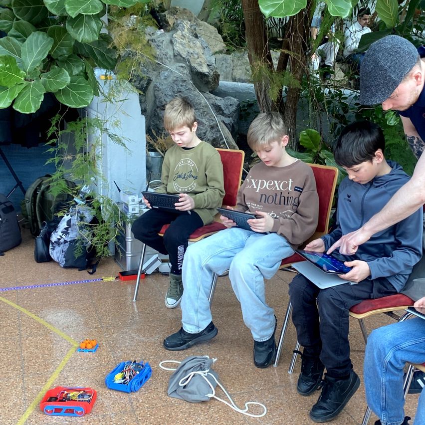 Vier Jungen sitzen auf Stühlen und schauen auf Tablets. Ein Mann in einer Mütze steht hinter ihnen und zeigt etwas auf dem Tablet.