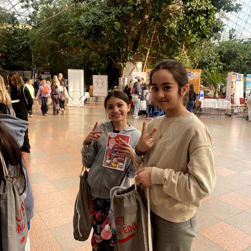 Zwei Mädchen posieren für ein Foto in einem Einkaufszentrum. Sie haben lächelnde Gesichter und machen Friedenszeichen.