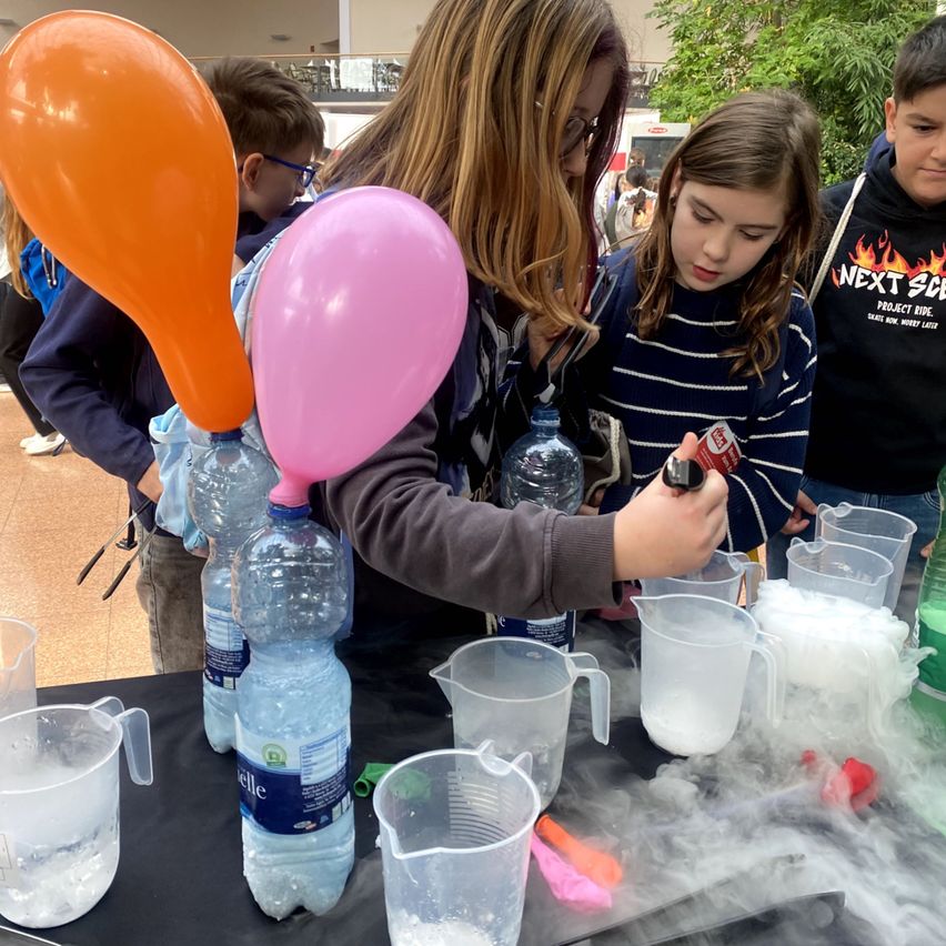 Mehrere Kinder stehen um einen Tisch herum und machen ein Wissenschaftsexperiment mit Wasser, Ballons und Messbechern.