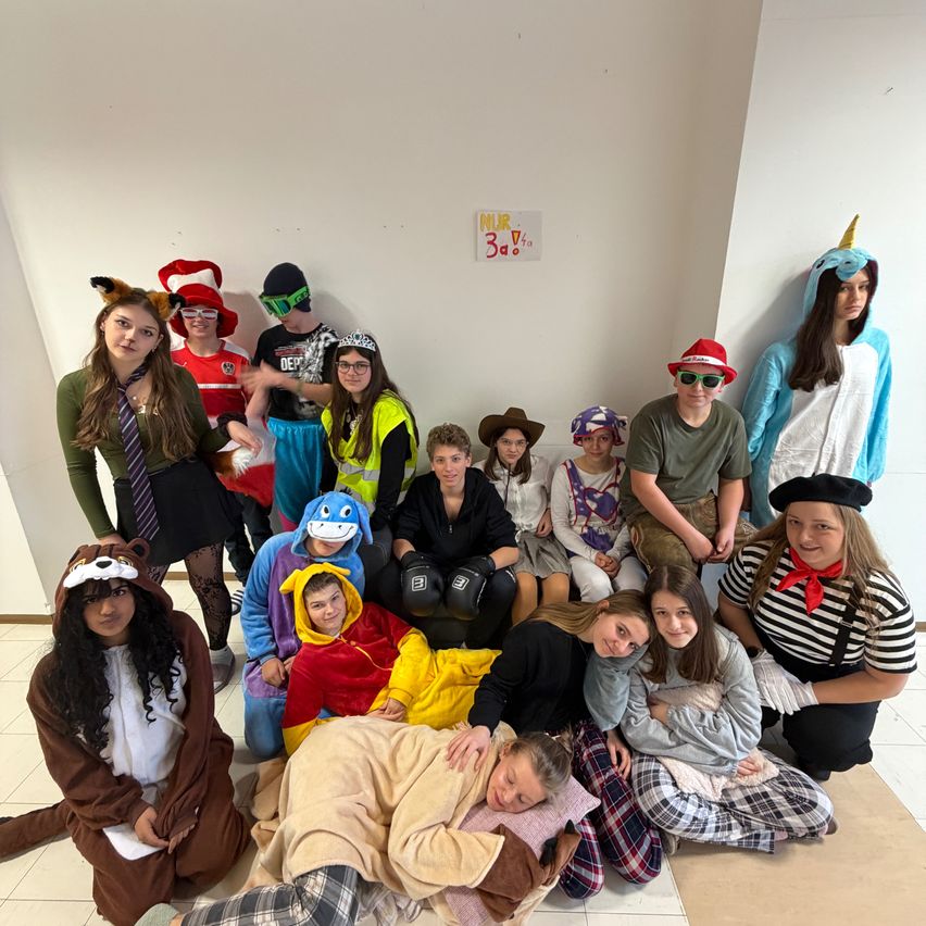 Eine Gruppe von Menschen in Kostümen posiert für ein Foto. Einige sitzen auf dem Boden, während andere hinter ihnen stehen. Der Raum hat weiße Wände und eine Tafel mit dem Text 'NLR 3d.V.'.