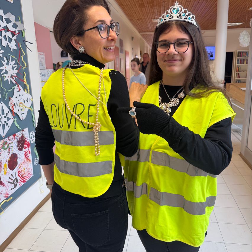 Zwei Frauen in Warnwesten stehen in einem Flur. Eine trägt eine Krone, und beide tragen eine Brille. Die linke Frau trägt eine Perlenkette. Hinter ihnen ist ein Mädchen in einem blauen Shirt zu sehen.