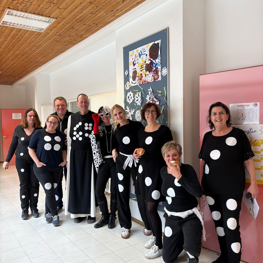 Eine Gruppe von Menschen in Tupfenkleidern steht in einem Flur, lächelt und posiert für ein Foto. Dahinter befindet sich eine Pinnwand mit Kunstwerken.