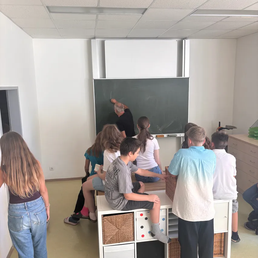 Ein Klassenzimmer mit einem Lehrer, der an eine Tafel schreibt, Schülern, die an Tischen sitzen, und einem weißen Schrank im Hintergrund.