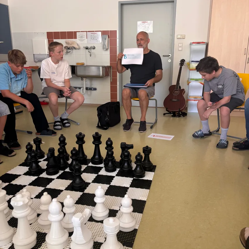 Eine Gruppe von Jungen und ein älterer Mann spielen Schach in einem Raum. Der ältere Mann hält ein Papier in der Hand und die Jungen sitzen auf Stühlen.