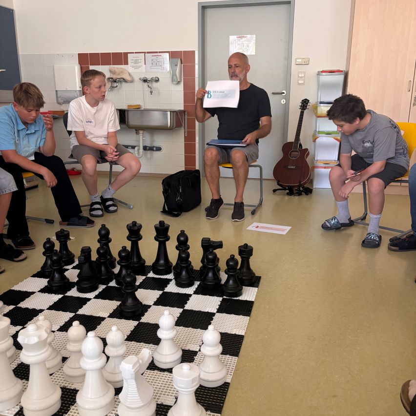 Eine Gruppe von Jungen und ein älterer Mann spielen Schach in einem Raum. Der ältere Mann hält ein Papier in der Hand und die Jungen sitzen auf Stühlen.