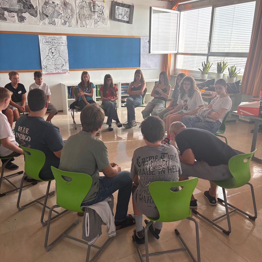 Eine Gruppe von Teenagern sitzt in einem Kreis auf grünen Stühlen, wahrscheinlich in einem Klassenzimmer. Einige unterhalten sich, während andere umherschauen. Der Raum hat eine blaue Wand mit Zeichnungen und eine Tafel.