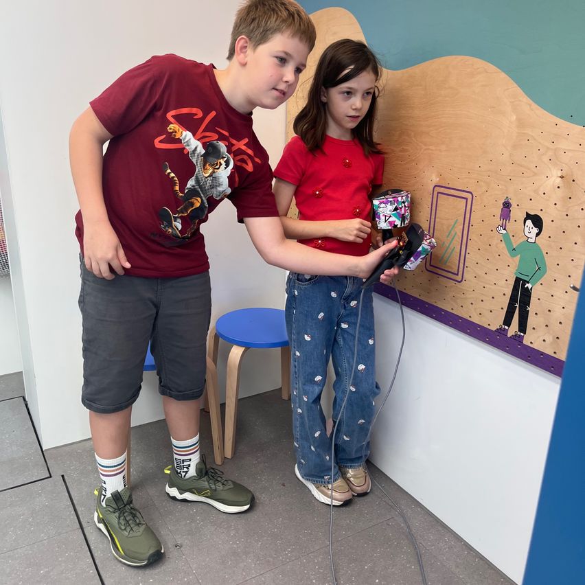 Zwei Kinder, ein Junge und ein Mädchen, stehen vor einer Wand mit einer Cartoon-Zeichnung und halten einen Spielcontroller.