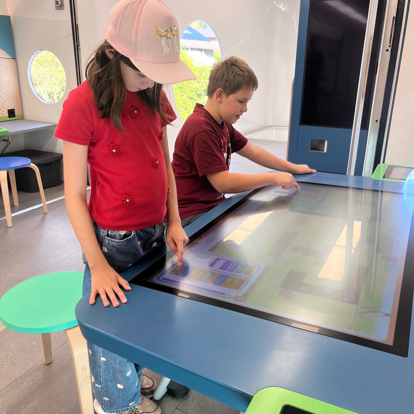 Zwei Kinder spielen auf einem großen interaktiven Bildschirm in einem Raum mit blauen Wänden und grünen Hockern.