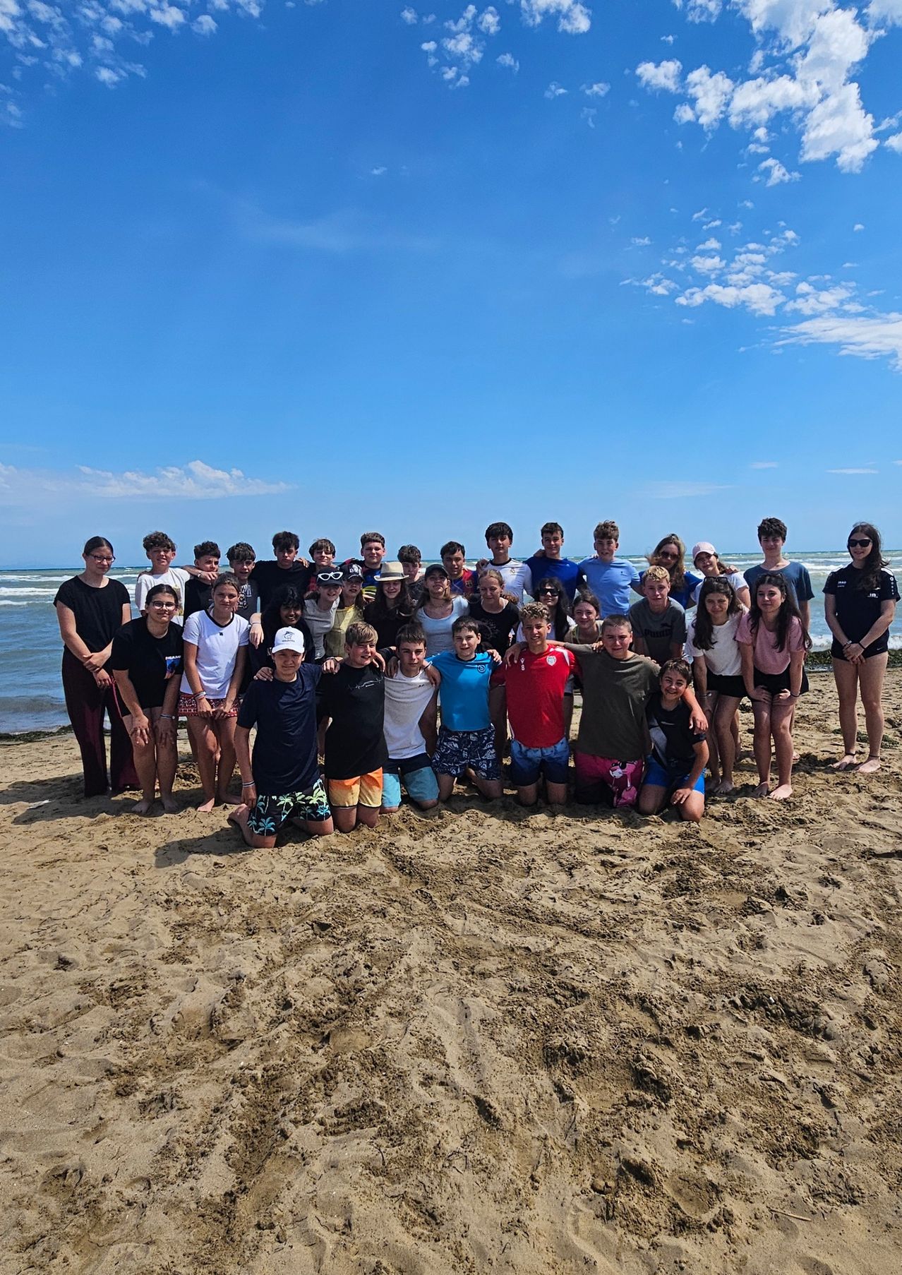 Eine Gruppe junger Menschen posiert für ein Foto am Strand, trägt Sommerkleidung und hat den Ozean und den Himmel im Hintergrund.
