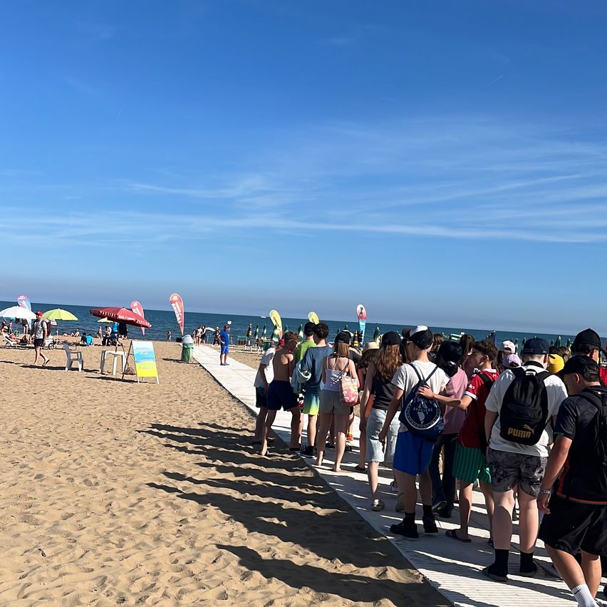 An einem sonnigen Tag warten eine Gruppe von Menschen mit Rucksäcken in einer Schlange auf einem Strandweg, mit Schirmen und einer Strandszene im Hintergrund.