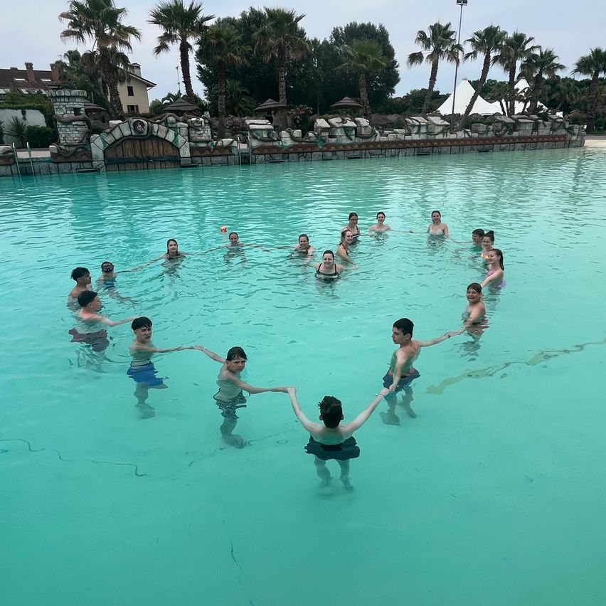 Eine Gruppe junger Menschen bildet ein Herz in einem Schwimmbecken an einem sonnigen Tag. Das Becken ist von Palmen und einer Steinmauer umgeben.