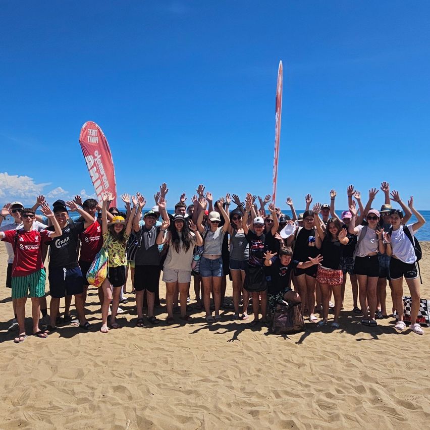 Eine Gruppe von Menschen posiert für ein Foto am Strand bei blauem Himmel. Einige tragen Sonnenbrillen und Hüte, während zwei Banner hinter ihnen stehen.