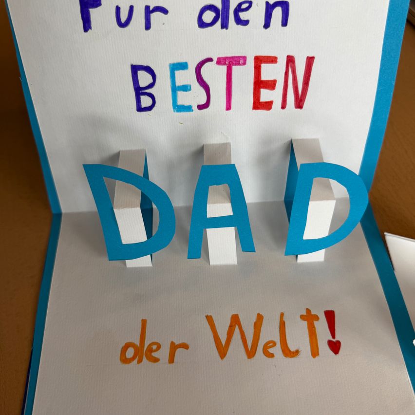 Eine handgemachte Karte mit Pop-up-Design trägt die Worte 'Für den besten Dad der Welt' in bunten Buchstaben. Die Karte hat einen weißen und blauen Rand.