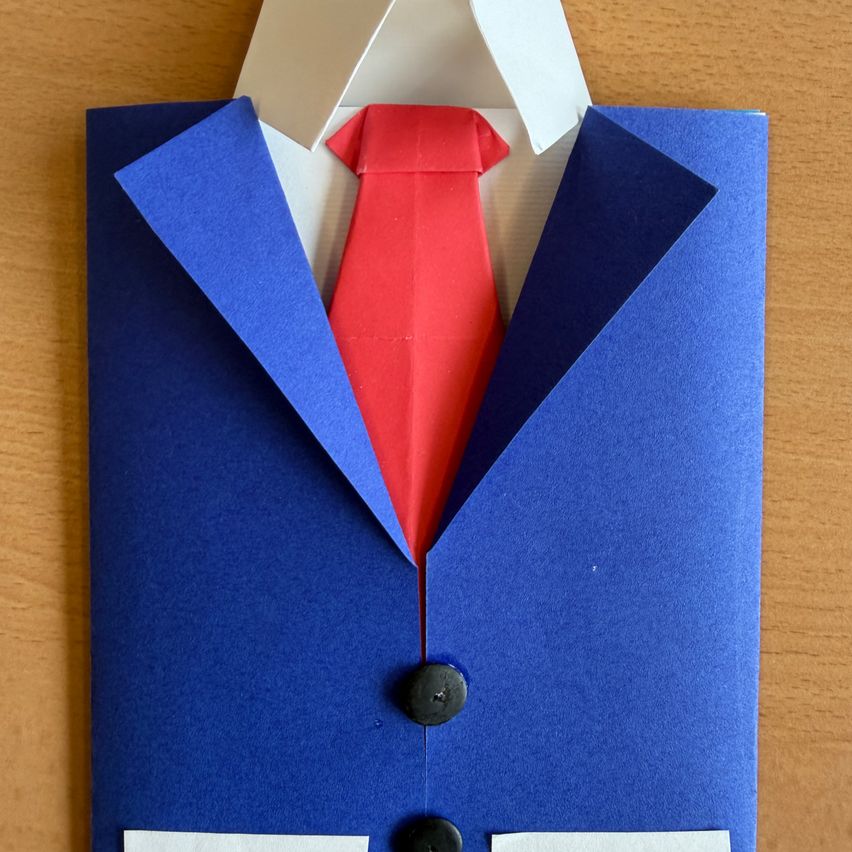 Ein blauer Papieranzug und ein weißes Hemd mit roter Papierkrawatte. Der Anzug hat einen Kragen und einen Knopf auf jeder Seite. Er liegt auf einer braunen Holzoberfläche.