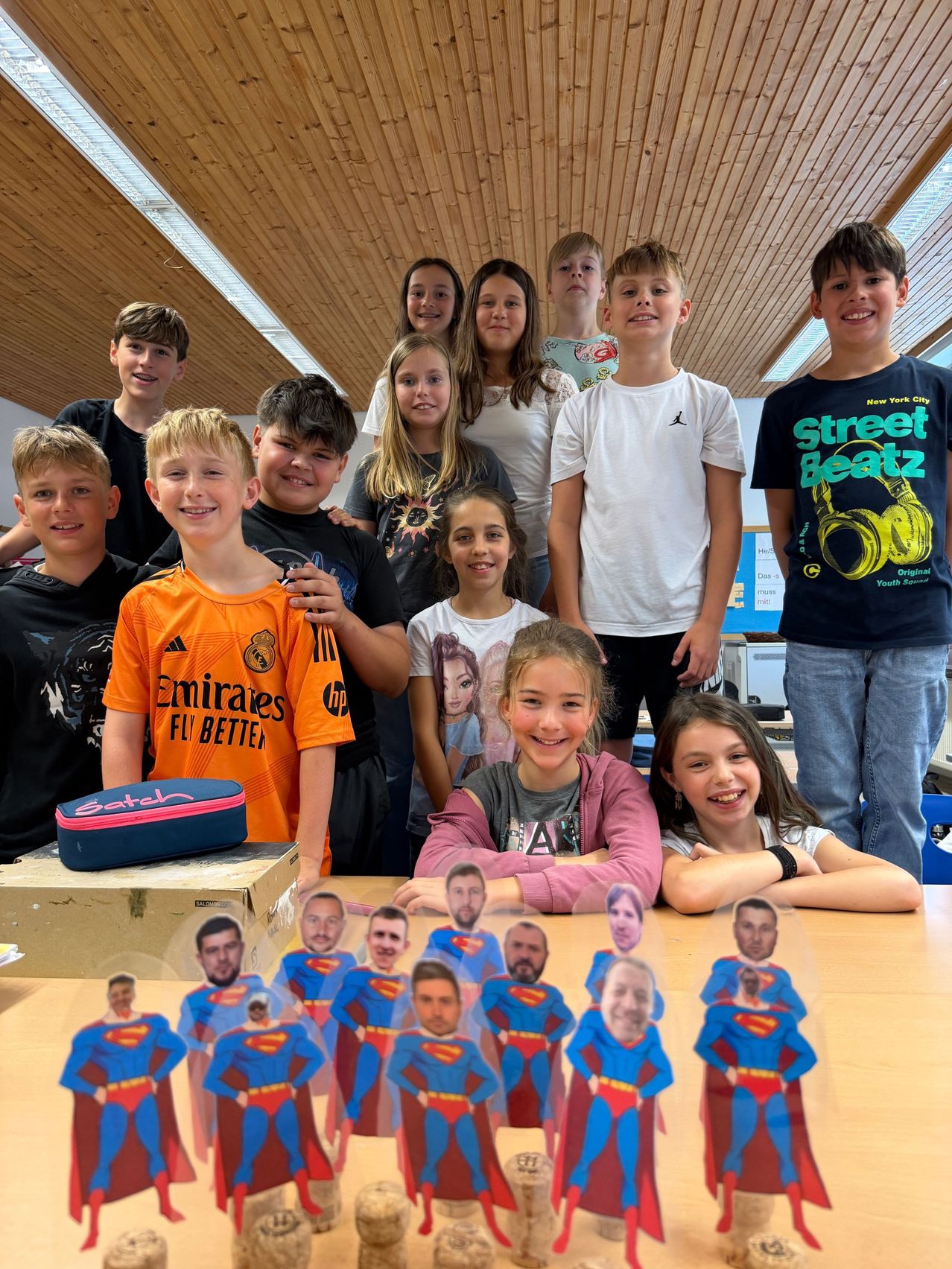 Eine Gruppe junger Menschen in einem Klassenzimmer, die lächelnd posieren und mit Superman-Ausschnitten auf einem Tisch.