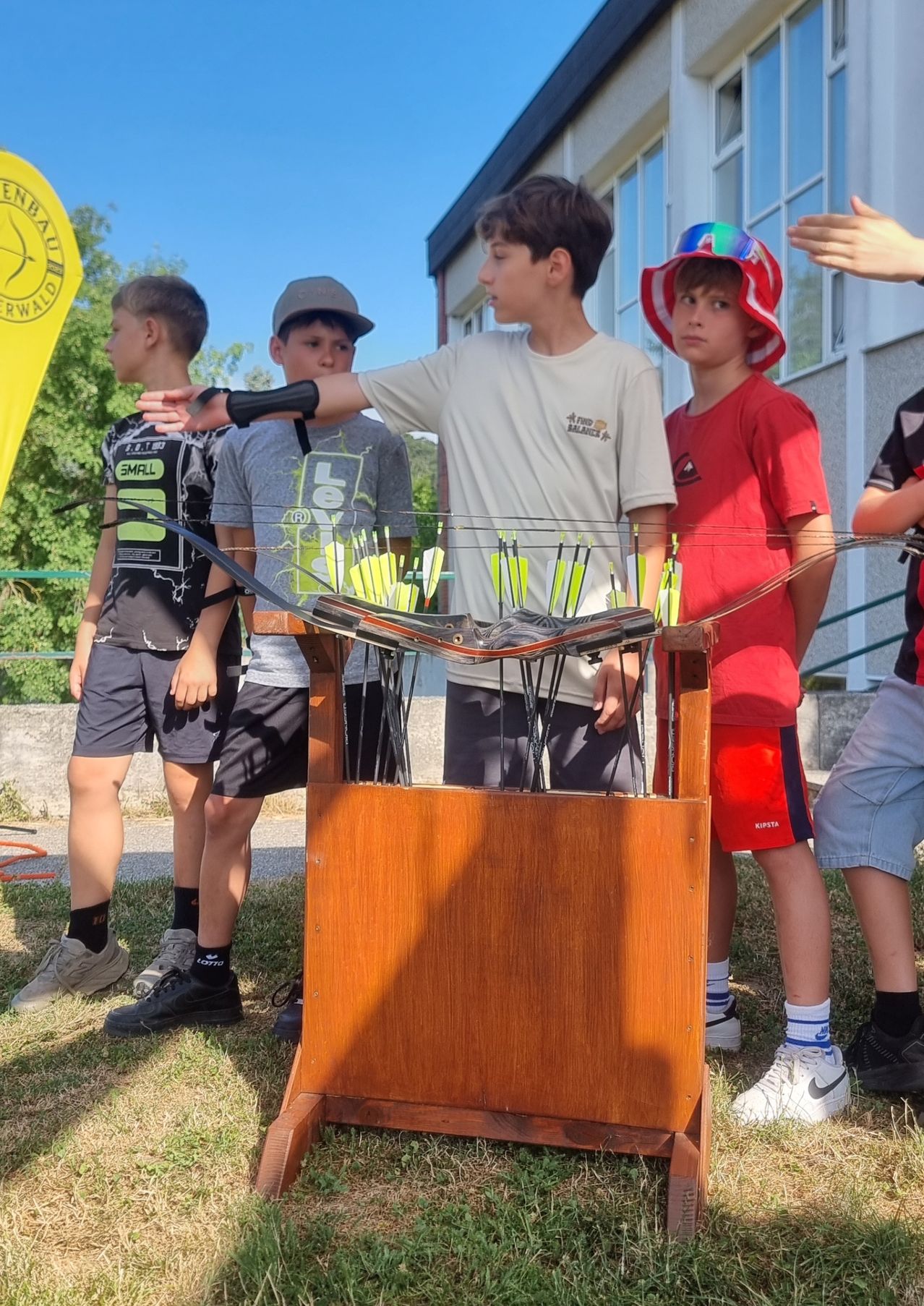 Five boys stand around a wooden archery target, holding bows and arrows. They seem to be participating in an archery event. A yellow flag is visible in the background.