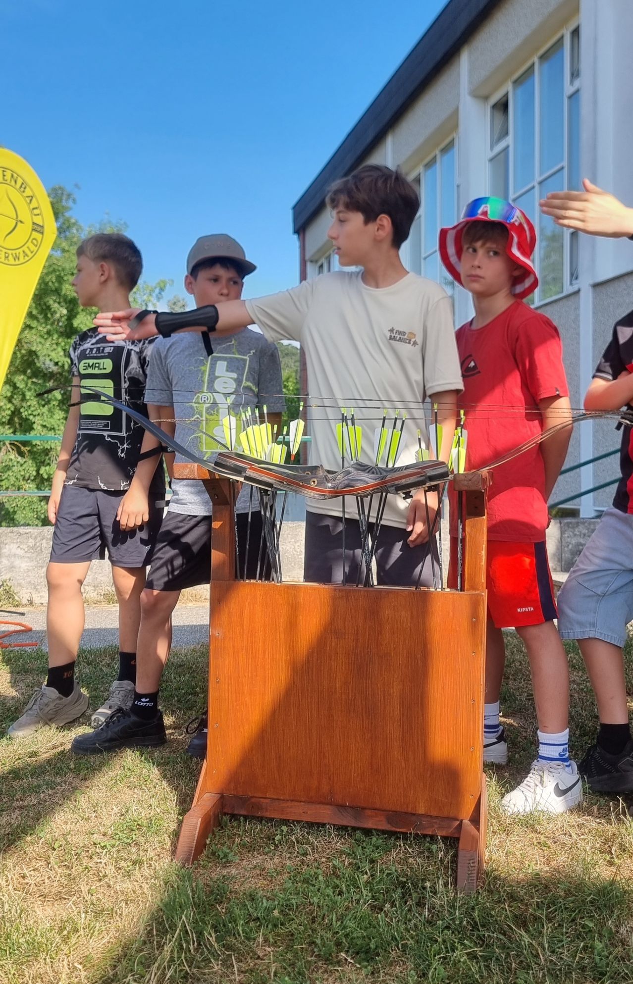 Fünf Jungen stehen um eine Holzpfeilwurf-Zielscheibe herum, halten Bögen und Pfeile. Sie scheinen an einer Bogenschießveranstaltung teilzunehmen. Im Hintergrund ist eine gelbe Fahne zu sehen.