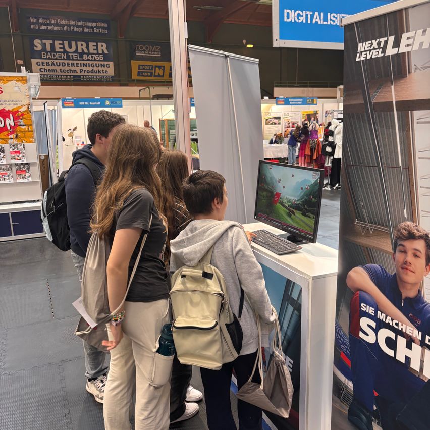 Eine Gruppe von Menschen versammelt sich um einen Computermonitor bei einer Veranstaltung, wobei eine Person aktiv ein Videospiel spielt. Der Schauplatz scheint eine Messe oder Ausstellung zu sein, mit verschiedenen Bannern und Werbeplakaten im Hintergrund.