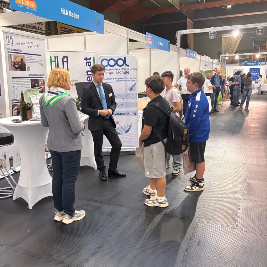 Eine Gruppe von Menschen versammelt sich bei einer Indoor-Veranstaltung, möglicherweise einer Messe. Ein Mann im Anzug spricht mit einer Frau in einer grauen Jacke. Dahinter ist ein Banner mit dem Wort 'COOL' zu sehen.