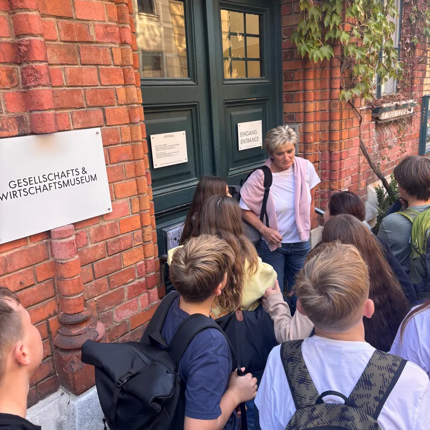 Eine Gruppe von Schülern, einige mit Rucksäcken, versammelt sich vor einem Backsteingebäude mit einem Schild, das Gesellschaft & Wirtschaftsmuseum besagt.