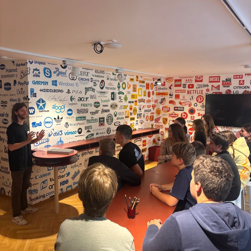 Eine Gruppe von Menschen sitzt um einen Tisch in einem Raum mit einer Wand voller Logos. Ein Mann steht vor ihnen und spricht.