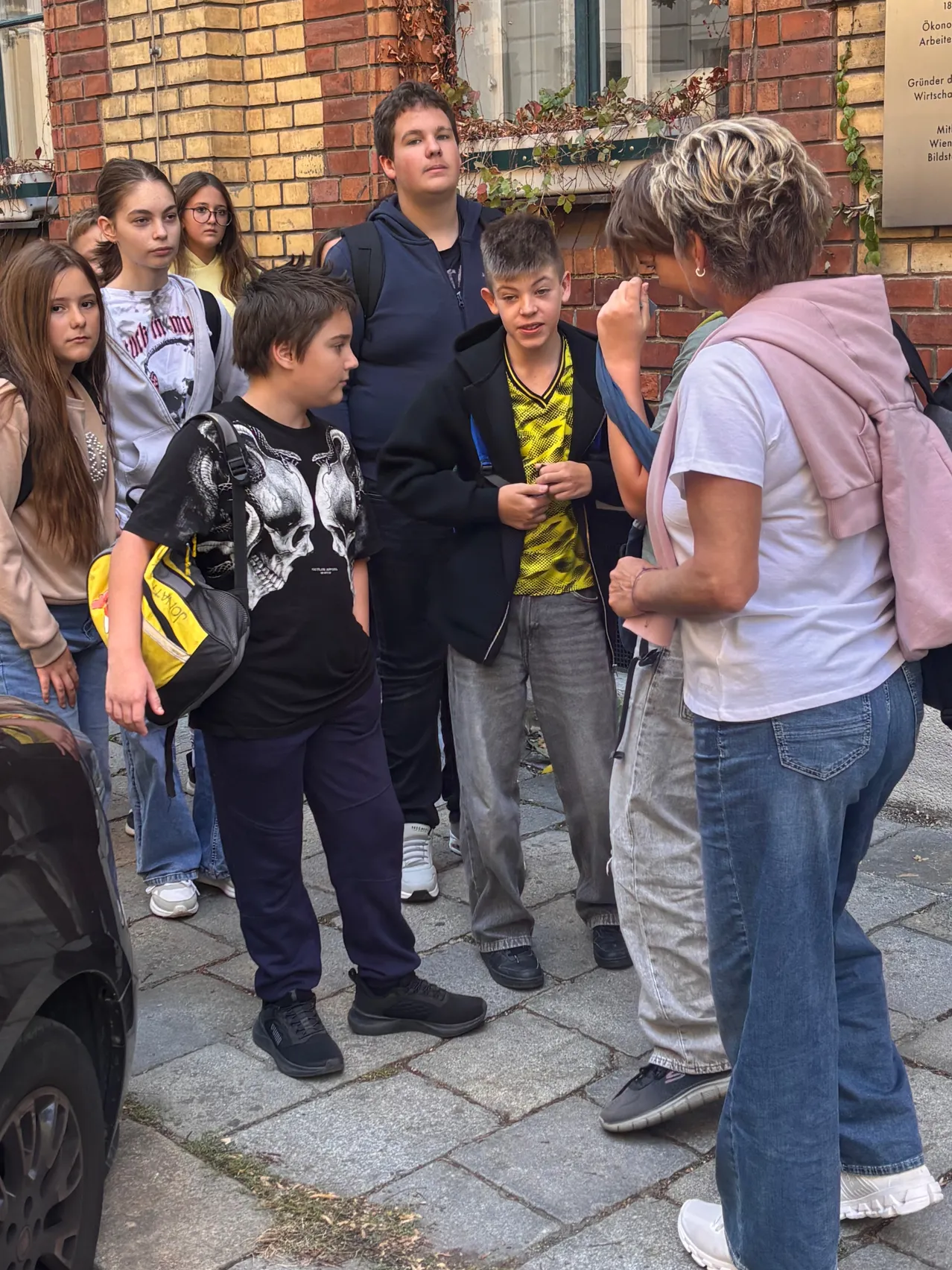 Eine Gruppe junger Leute steht vor einem Backsteingebäude. Eine Frau spricht mit einem Jungen, der ein Totenkopfhemd trägt.