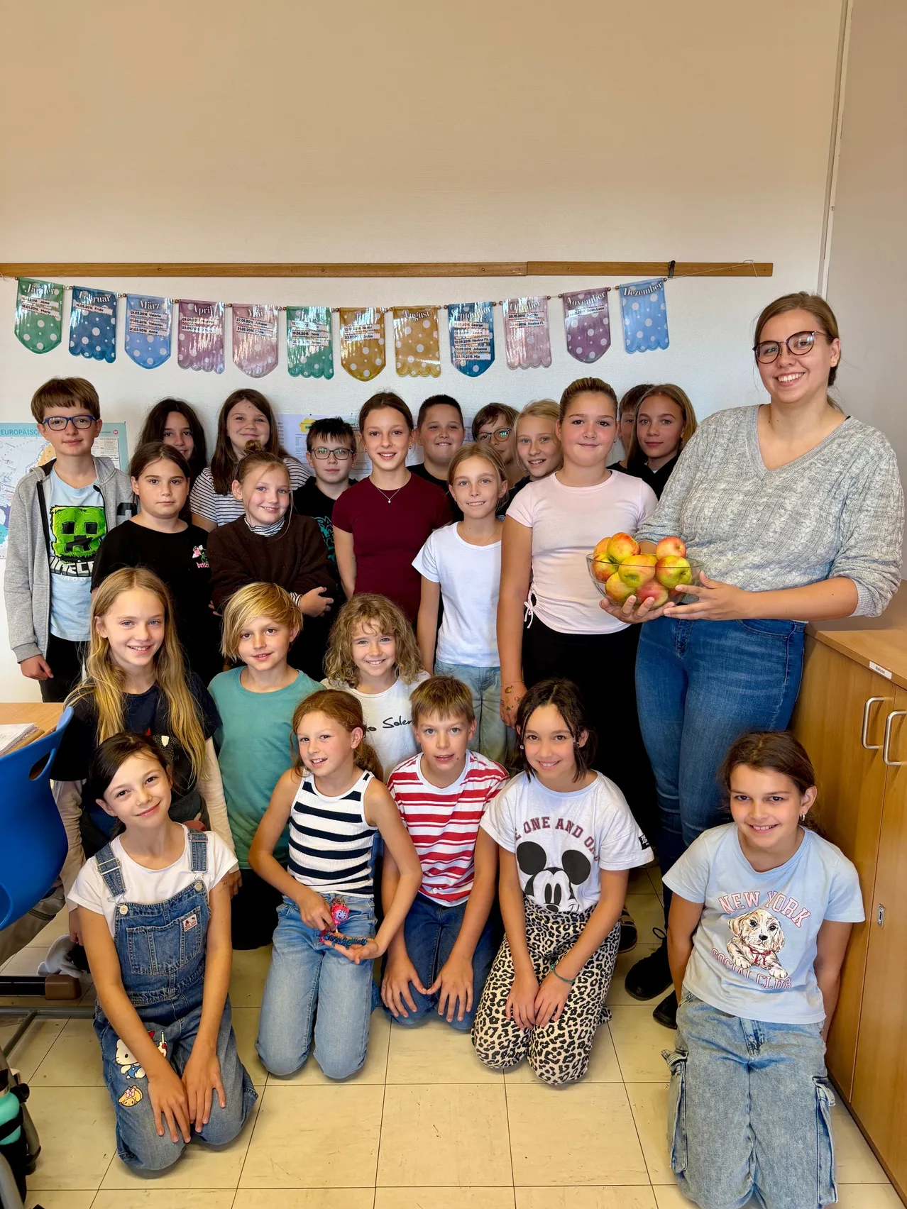 Eine Gruppe von Kindern und einer Frau posiert für ein Bild in einem Klassenzimmer. Die Frau hält eine Schüssel mit Äpfeln.