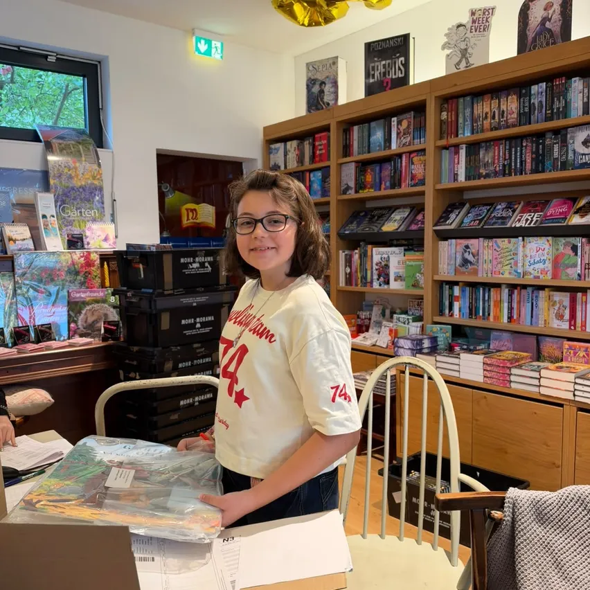 Ein junges Mädchen mit einer Brille steht in einem mit Büchern gefüllten Raum und hält eine Schachtel und einen Plastikbeutel.