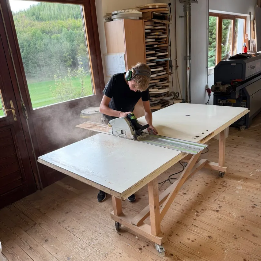 Ein Mann in einem schwarzen Shirt und Kopfhörern benutzt eine Kreissäge, um ein Stück Holz auf einem Holztisch in einer Werkstatt zu schneiden.