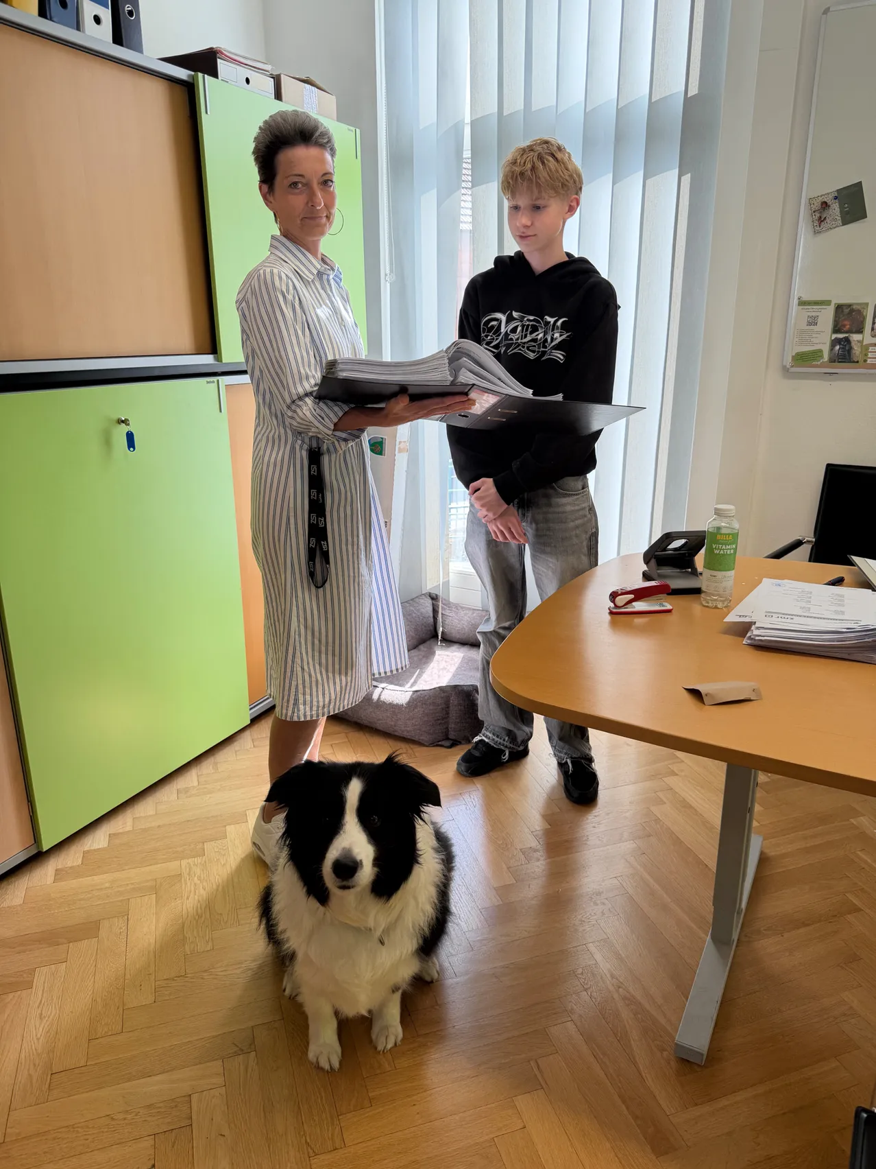 Eine Frau und ein junger Junge stehen in einem Büro. Die Frau hält eine Mappe und einen Stift, und der Junge hält eine Mappe. Ein Hund sitzt neben ihnen auf dem Boden.
