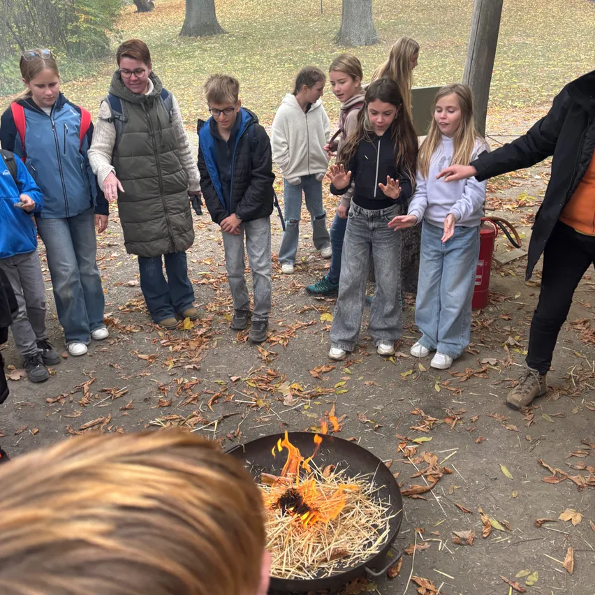Eine Gruppe von Kindern und Erwachsenen versammelt sich um ein Feuerloch im Freien. Das Feuerloch brennt mit Heu, und die Kinder beobachten es.