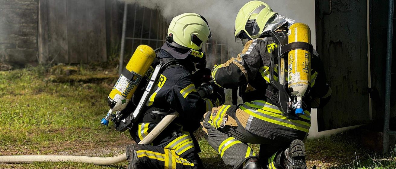 Zwei Feuerwehrleute in voller Ausrüstung knien auf Gras, einer sprüht Wasser, während der andere den rauchgefüllten Bereich überwacht.