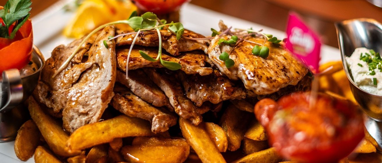 Ein Gericht mit verschiedenen Fleischsorten, Kartoffeln und Gemüse auf einem weißen Teller. Eine Wurst, Tomate und Petersilie obenauf, mit Beilagen auf der Seite.