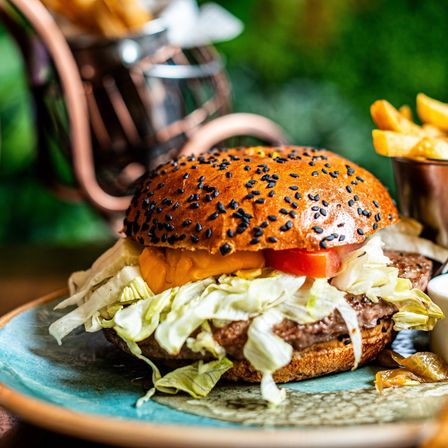 Ein Hamburger mit Sesamkörnern, Salat, Tomate und Käse auf einem blauen Teller mit Pommes.