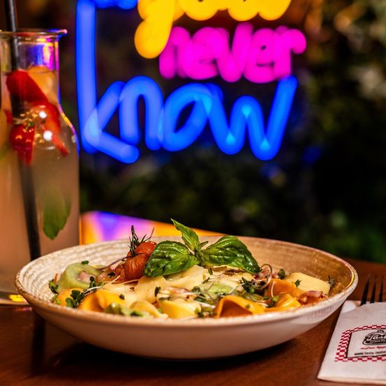 Eine Schüssel mit Nudeln und Gemüse, ein Glas Wasser mit einer Erdbeergarnitur und ein Neonschild, das 'Du weißt nie' besagt.