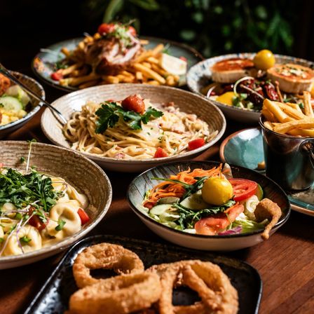 Ein hölzerner Tisch bietet verschiedene Gerichte: Pasta, Salate und frittierte Speisen. Jedes Gericht wird in seinem eigenen Teller oder Teller serviert. Pommes sind in einer Metalltasse.