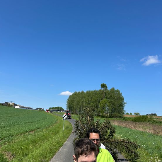 Zwei Männer gehen auf einem Pfad, der von grünem Gras umgeben ist. Einer trägt eine Brille und ein grünes Shirt. Der andere trägt ein weißes Shirt. In der Ferne fährt ein Traktor.