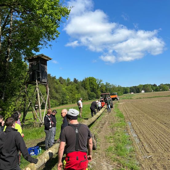 Eine Gruppe von Menschen steht auf einem Grasfeld, in der Ferne sind Traktoren geparkt. Eine Person hält einen Holzstamm.