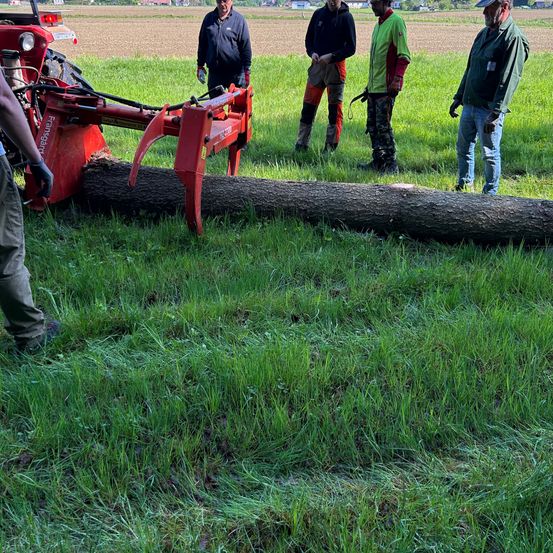 Fünf Männer stehen auf einem Grasfeld, einer hält eine rote Maschine. Der Mann vorne bedient die Maschine. Dahinter steht ein Traktor mit Anhänger.