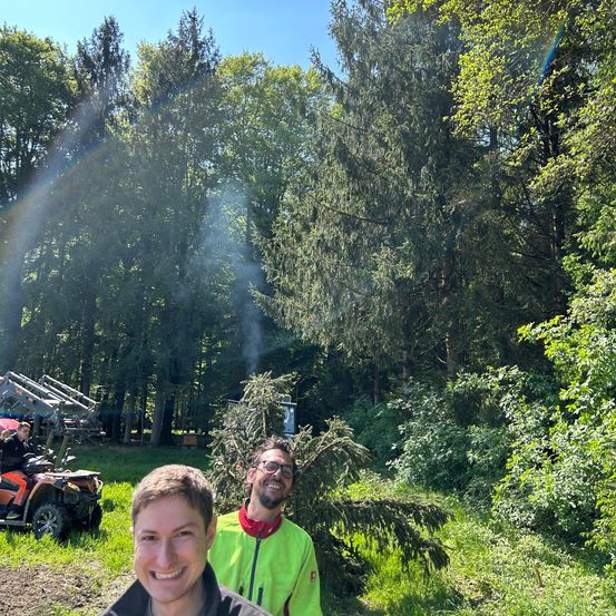 Zwei Männer stehen in einem Wald. Einer trägt eine neongrüne Jacke, der andere hat ein strahlendes Lächeln. Hinter ihnen liegt ein umgestürzter Baum auf dem Boden. Im Hintergrund ist ein Fahrzeug geparkt, und das Sonnenlicht erzeugt einen Regenbogeneffekt.