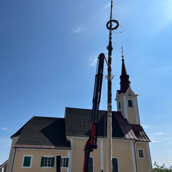 Ein Kran hebt einen dekorativen Pfahl vor einer Kirche mit einem Kirchturm und Uhrenturm.