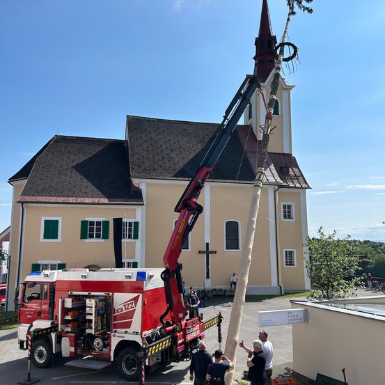 Ein Feuerwehrauto mit einem Kran hebt einen großen Baum vor einer Kirche.