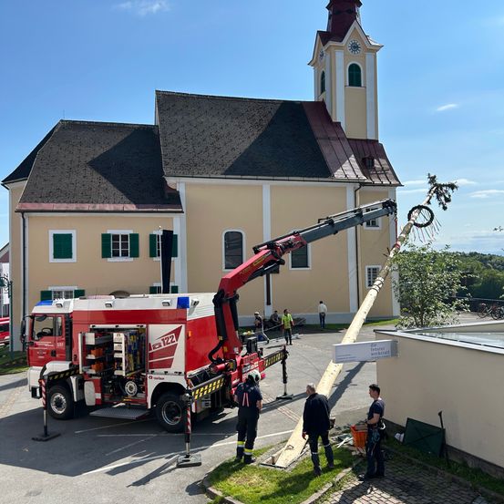 Ein Feuerwehrwagen ist vor einer Kirche geparkt. Ein langer Holzbalken wird von einem Kran angehoben. Vier Personen stehen neben dem Balken, von denen eine einen Schutzhelm trägt.